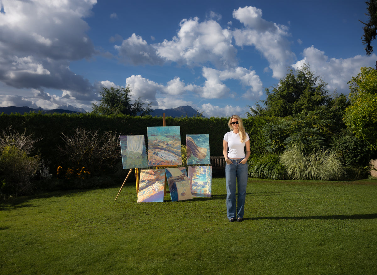 A woman stands on a green lawn outdoors next to several landscape paintings displayed on easels, with trees, a blue sky, and mountains in the background.