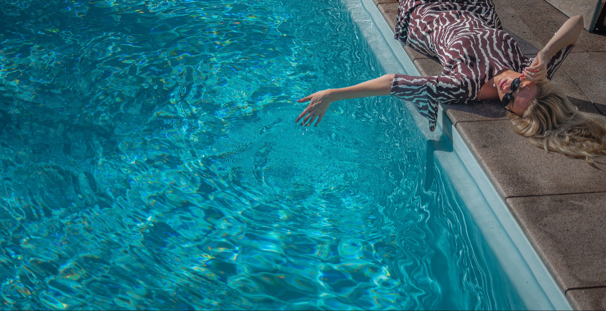 A woman wearing a brown and white zebra-print swimsuit lies on her back on the edge of a swimming pool, with her arm reaching into the bright blue water.