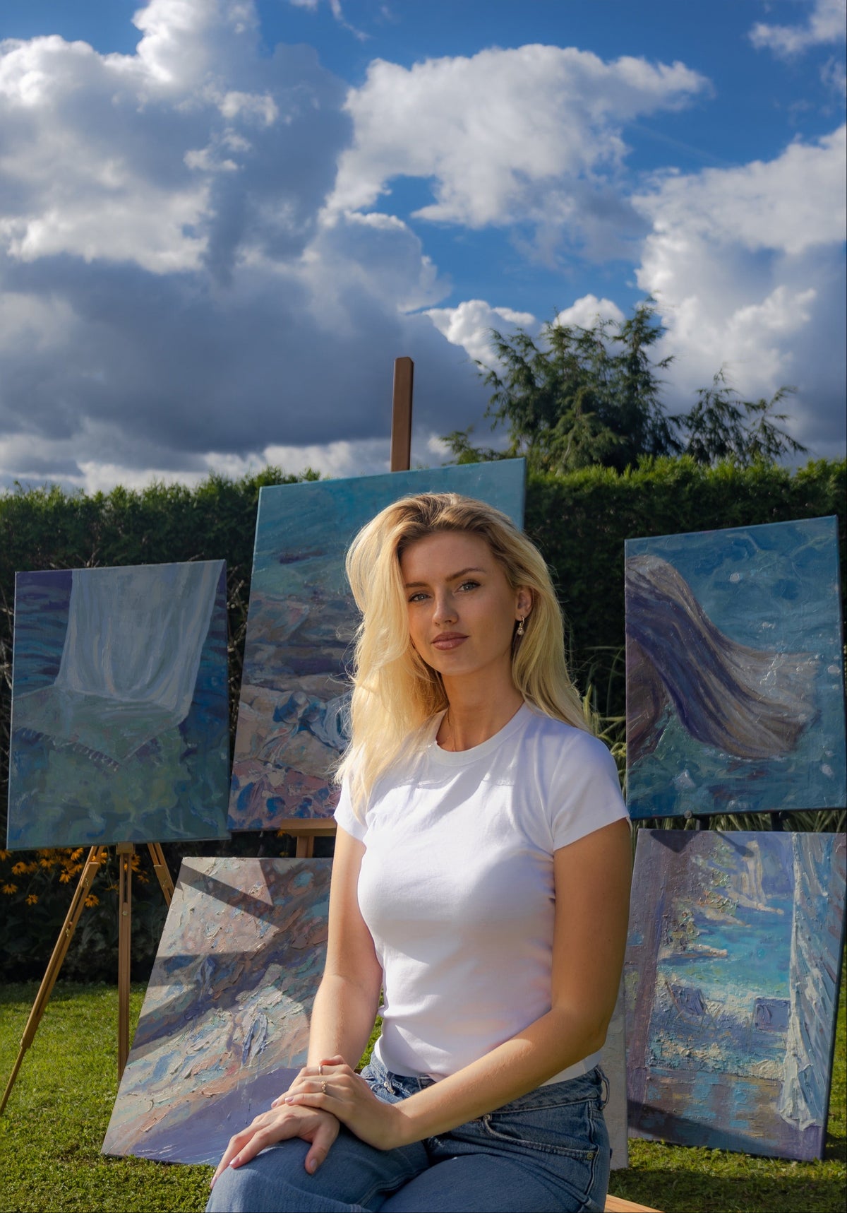 A woman with blonde hair sits on a lawn in an outdoor garden setting, wearing a white t-shirt and blue jeans. She is positioned next to several canvas paintings on easels, with a green hedge and a blue sky filled with white clouds behind her.